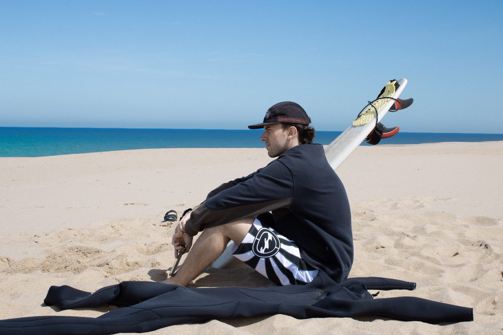 Surfista sentado en la arena con su tabla y un gorro Shakas mirando el mar