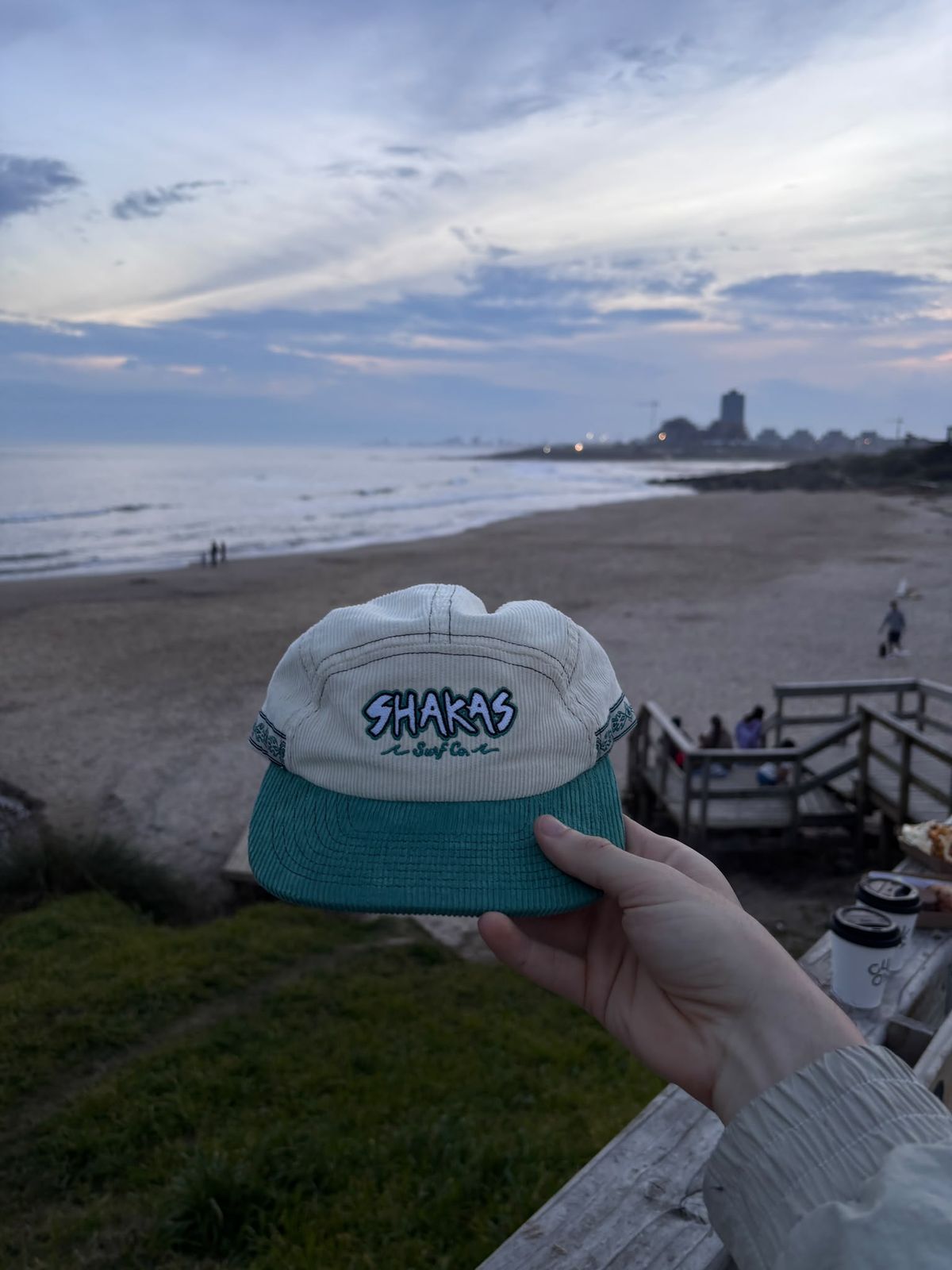 Mano sosteniendo el gorro Salty Hair frente a la playa al atardecer